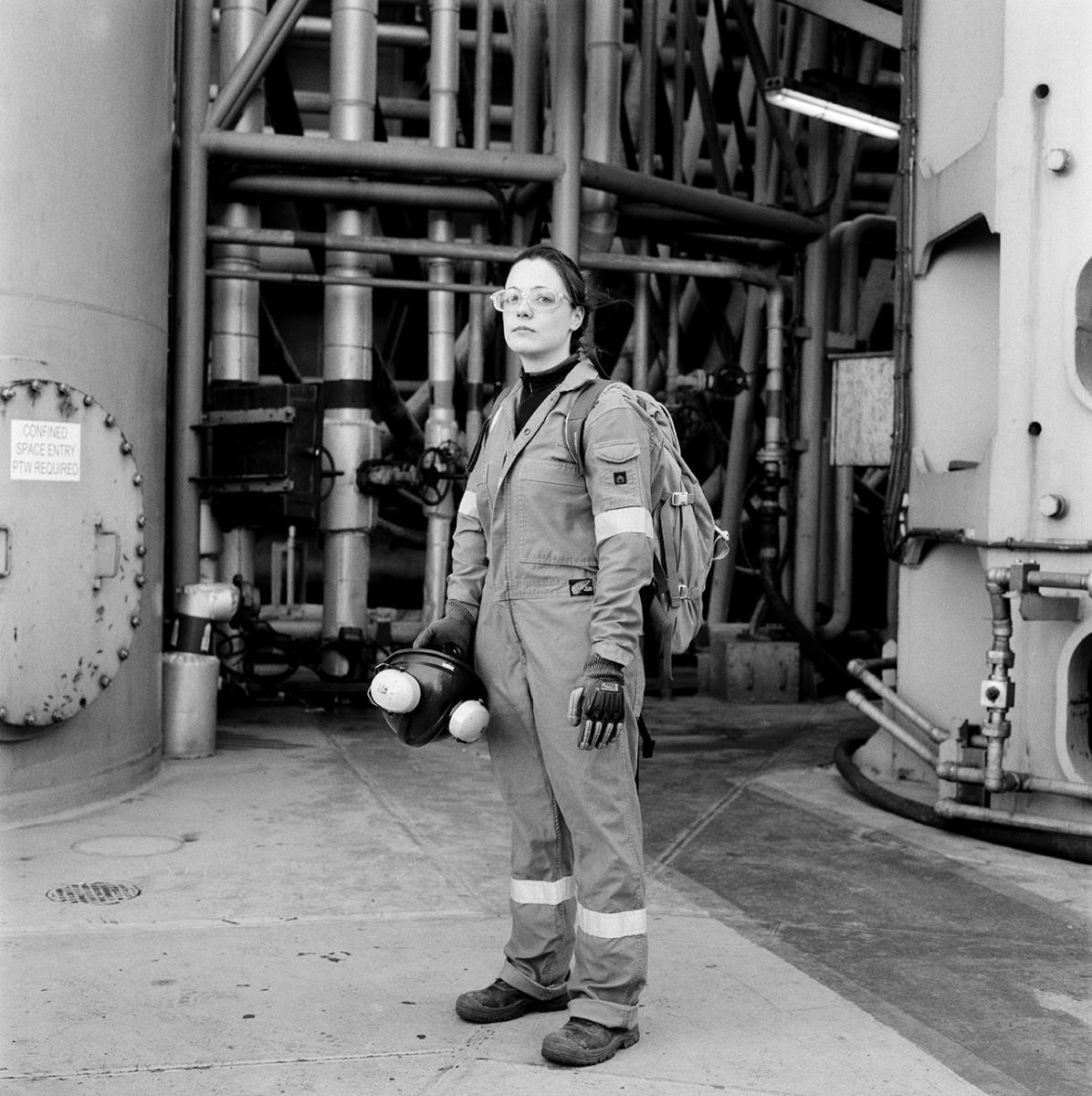 A black and white photo of an oil worker. She wears glasses and overalls, and stands looking at the camera in front of rig pipes