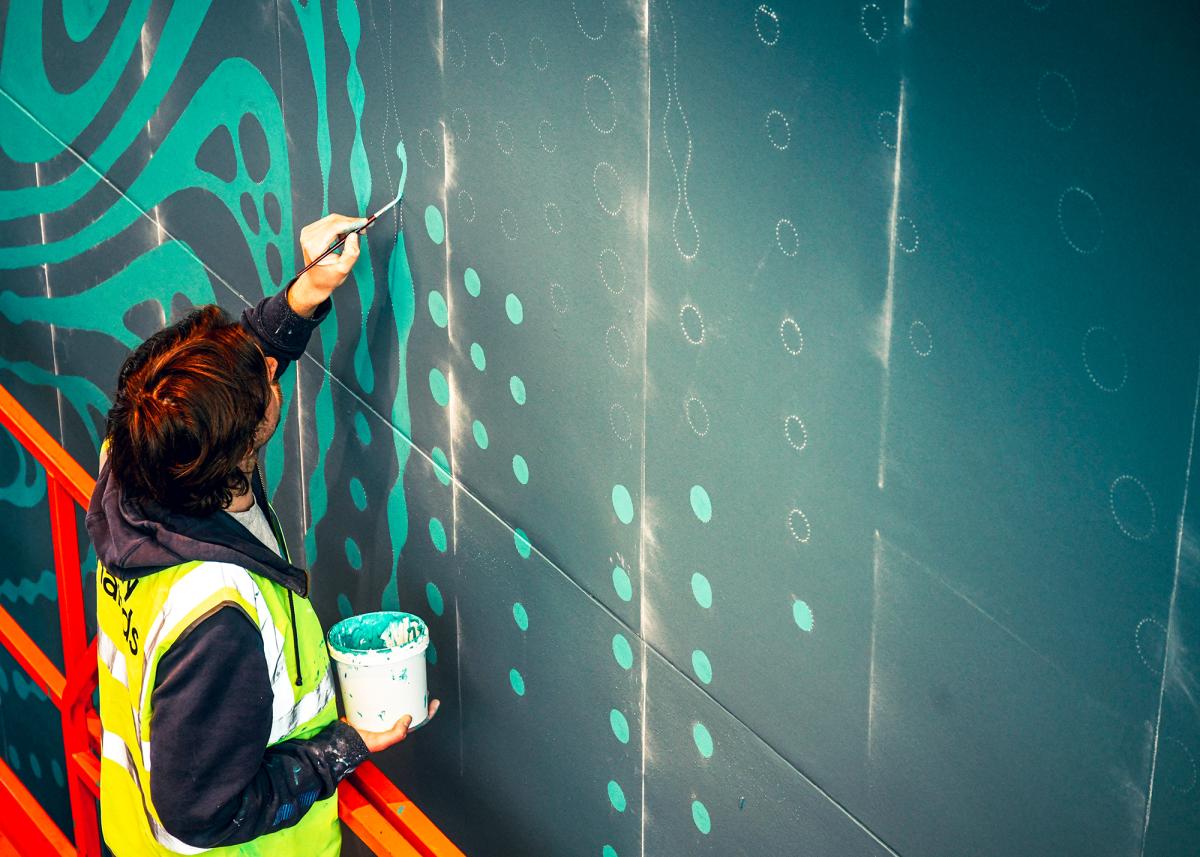 A man in a yellow high-vis jacket works on a swirling blue-green mural design on the wall of the National Maritime Museum.