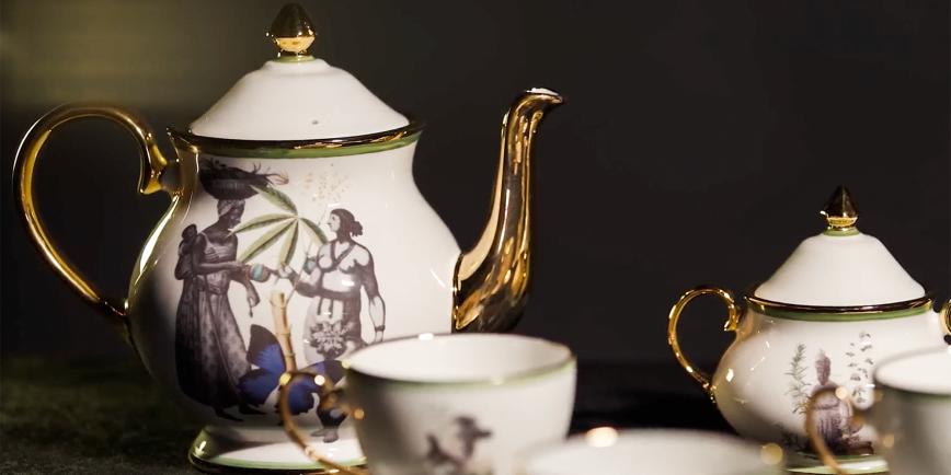 A delicate-looking tea service featuring teapot and matching cups. The central motif features a Black woman with a child on her back, handing an item to a bare-chested figure