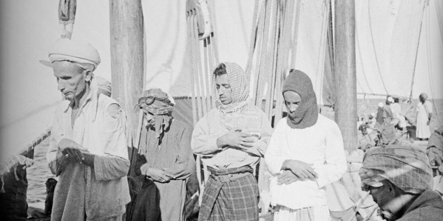 Historic black and white photograph of men  on board ship wearing head coverings and bowing their heads in prayer