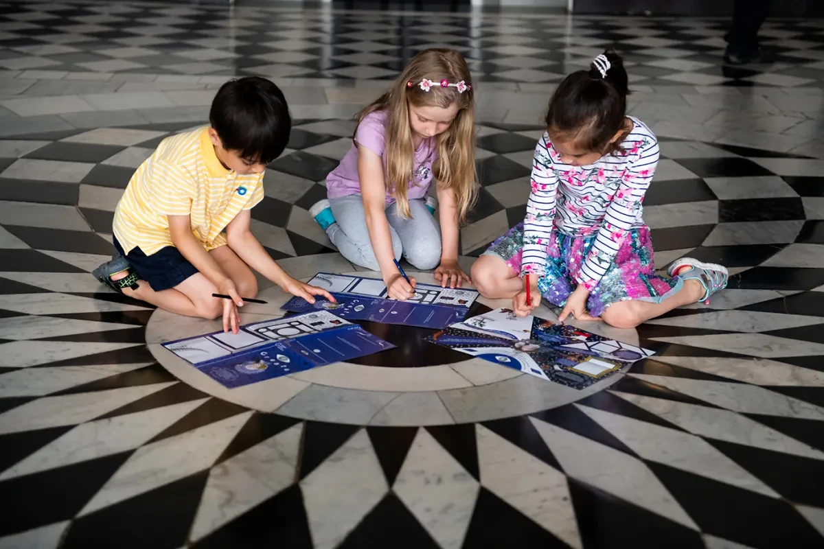 Children fill out an activity sheet during a visit to the Queen's House in Greenwich. They are sat on the floor, at the centre of an intricate black and white marble pattern