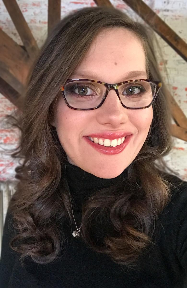 A woman with dark brown wavy hair and glasses smiles at the camera