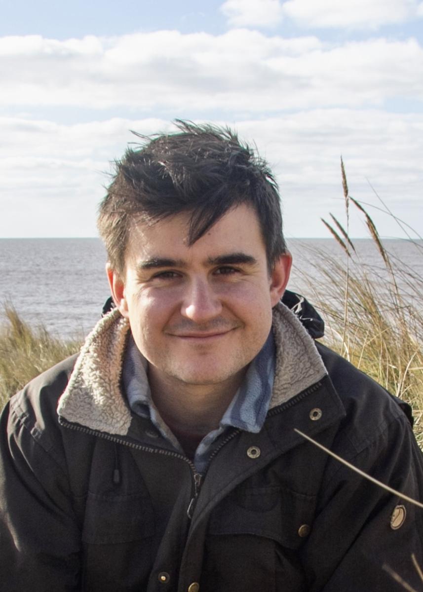 A man wearing a fleece-lined outdoor jacket smiles at the camera, with a view of the sea and grass behind