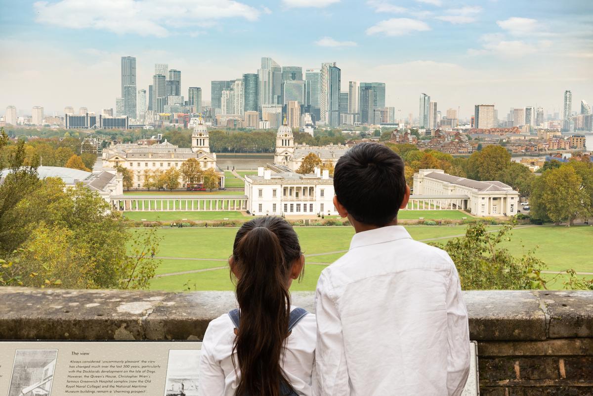 A brother and sister with their back to the camera look out from the viewpoint at the top of Greenwich Park towards historic Greenwich