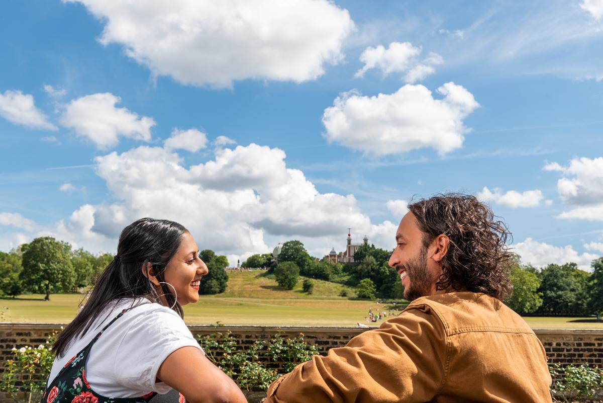 A couple sits on a bench with Greenwich Park and the Royal Observatory in the background