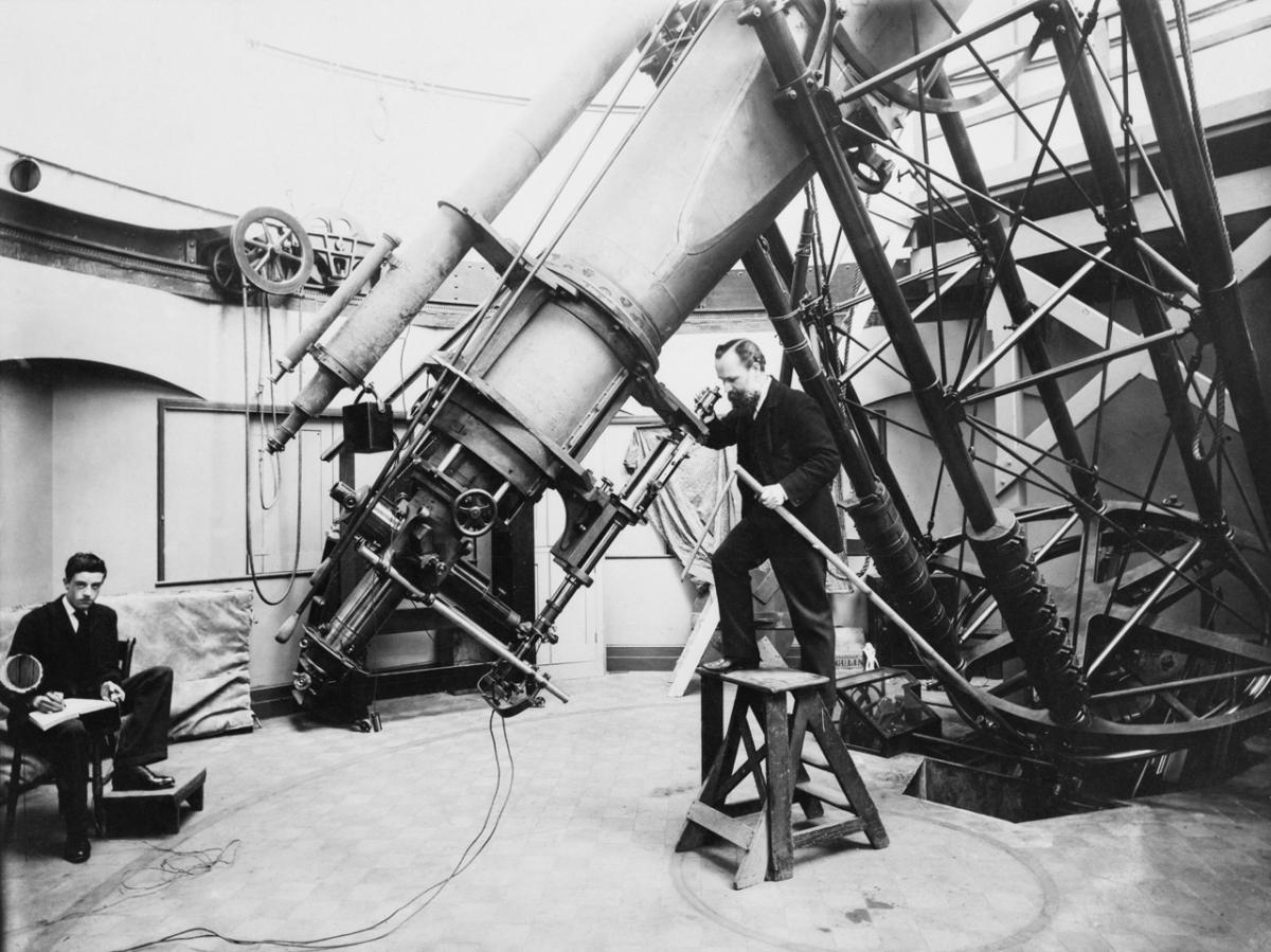 Historic photograph showing an astronomer peering into the eyepiece of the Great Equatorial Telescope at the Royal Observatory. Another astronomer is taking notes to the left