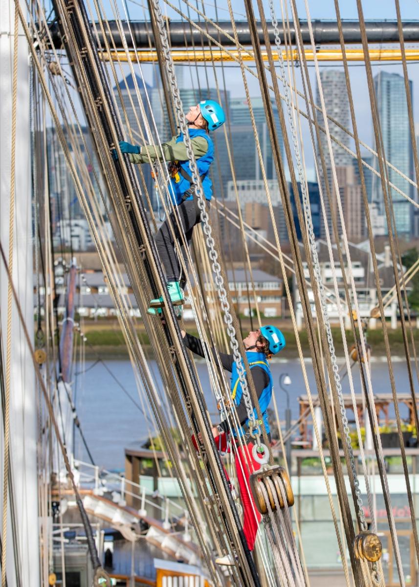 Two people climb the rigging of Cutty Sark, with views of London's Canary Wharf skyline in the background