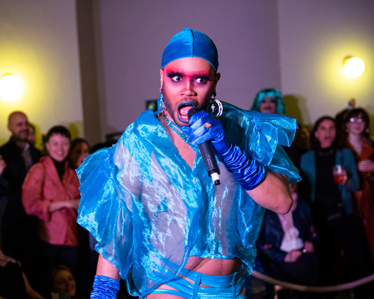 A drag performer wearing a blue swimming cap, velvet gloves and blue silk shirt