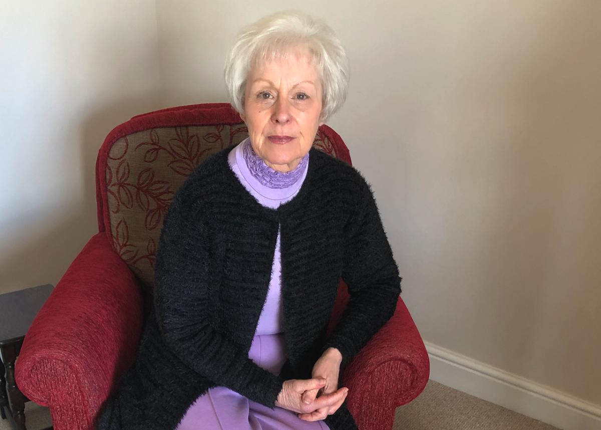 A photograph of Liz Ormerod, who served on the SS Uganda hospital ship during the Falklands conflict. She is sitting in a red chair looking straight at the camera, wearing a dark cardigan and light purple dress