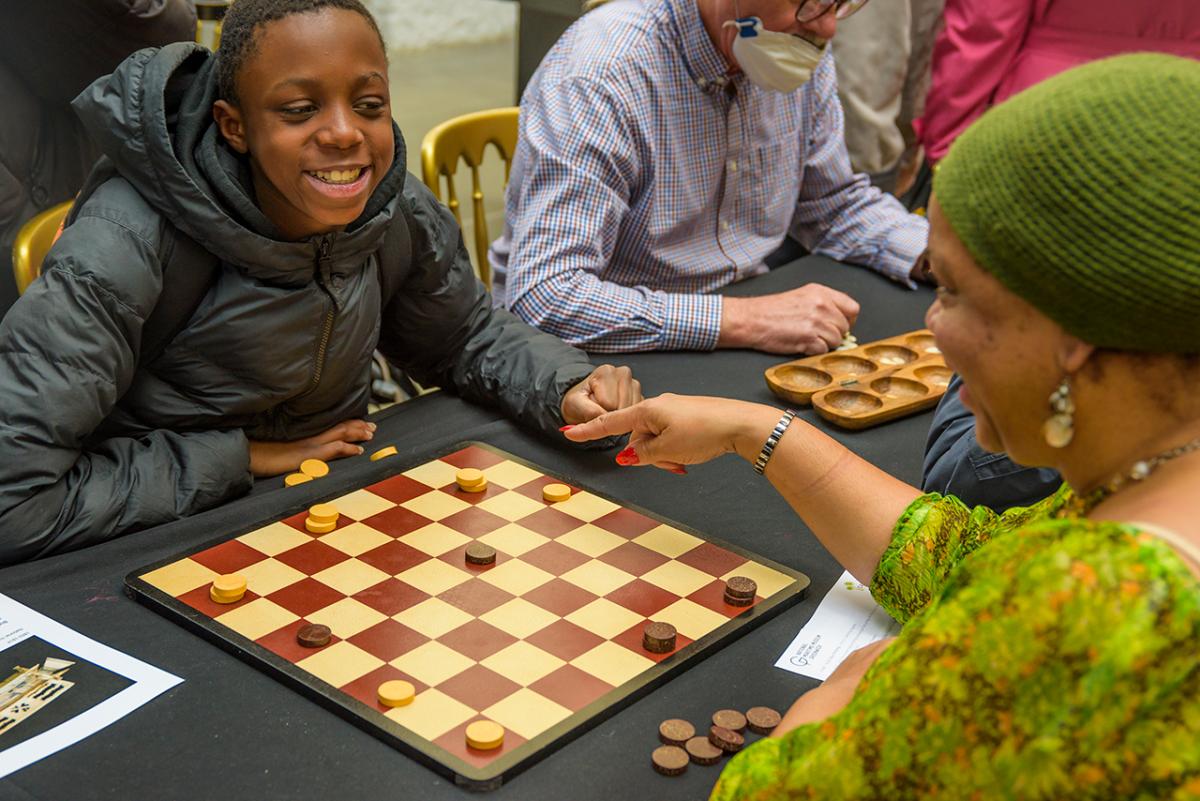 A boy smiles as he plays a game of draughts with a woman in a bright green dress