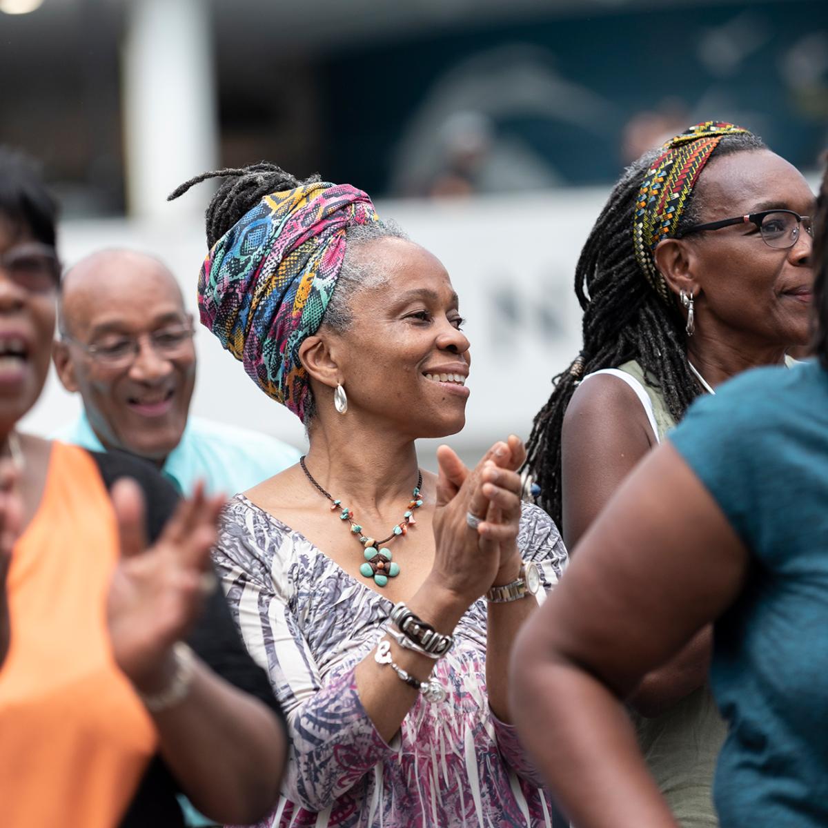 A woman wearing a brightly coloured headscarf smiles and claps during a musical performance