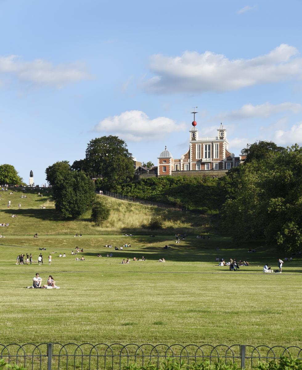 A view of Greenwich Park in summer, looking up the hill towards the Royal Observatory. People are sat enjoying the sunshine in the park