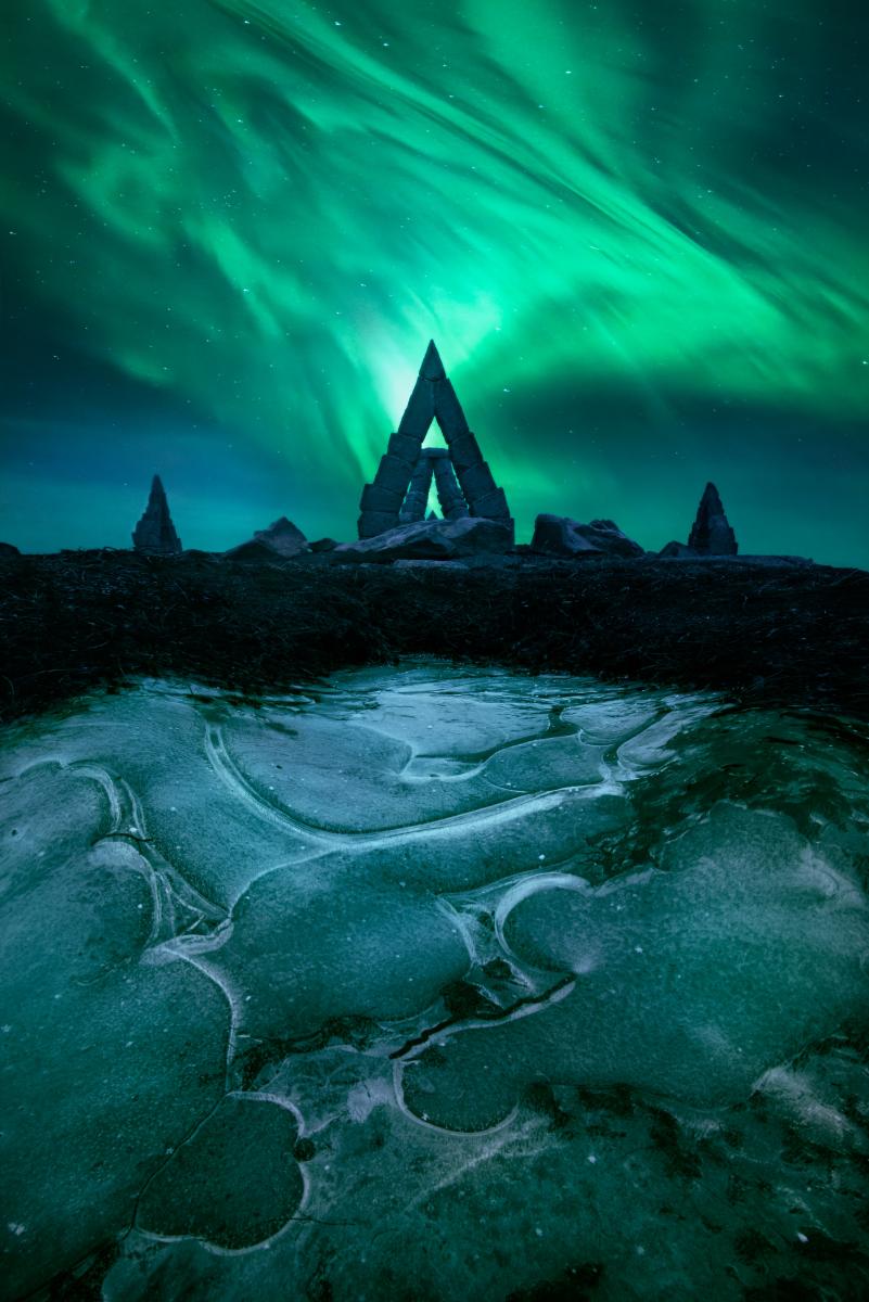 Image of aurorae swirling behind a stone gate in a triangular shape, with frozen water in the foreground