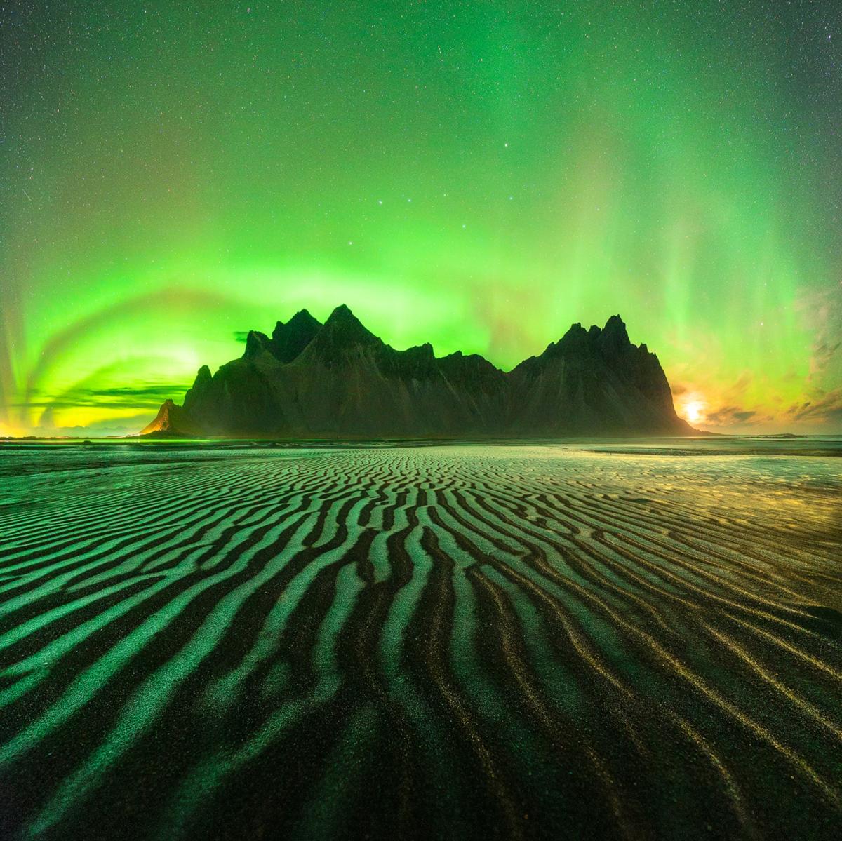 Image of a mountain with green aurorae in the sky behind it and reflected on the sand in the foreground