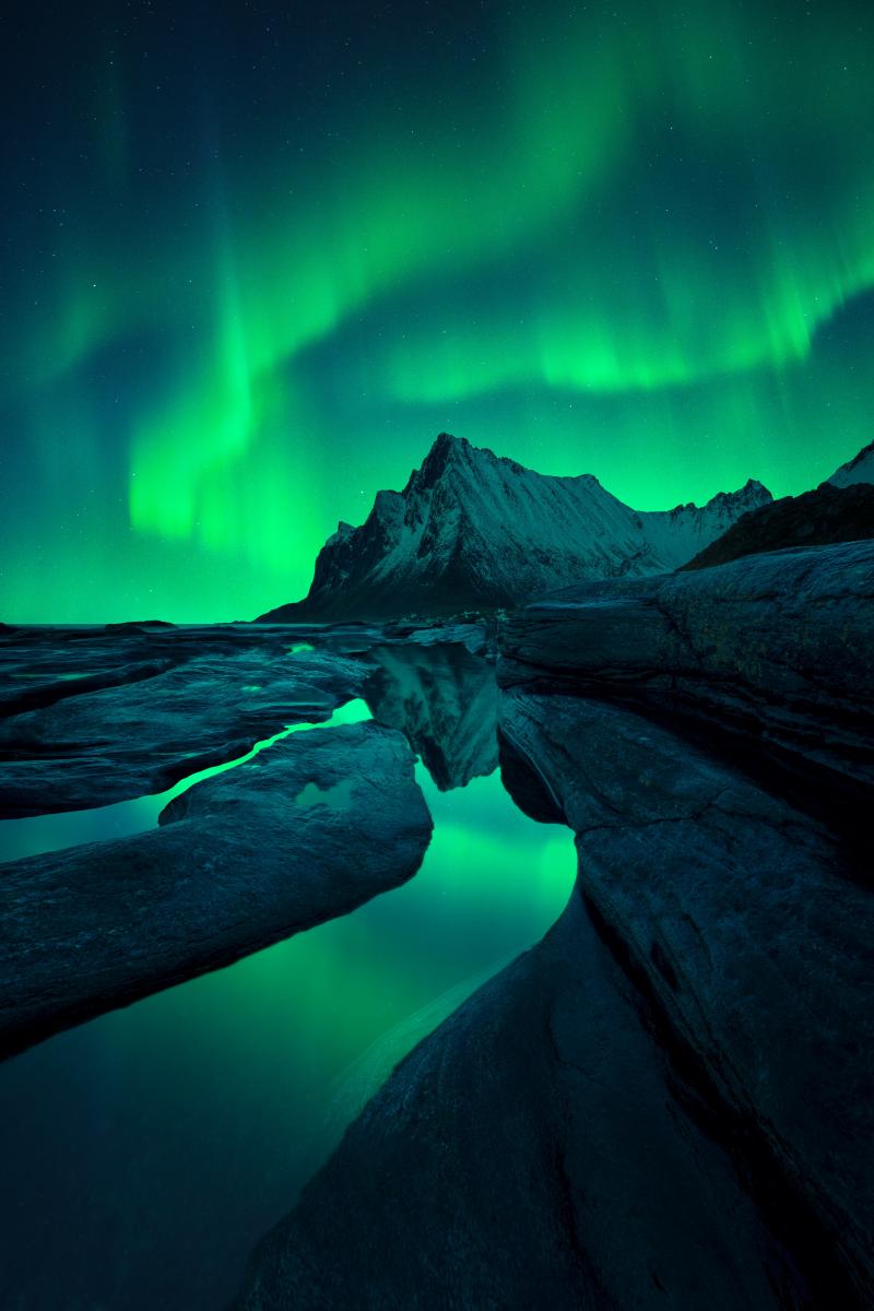 Image showing mountain and rocks in foreground, with rippling emerald aurora in the sky behind the mountain, which is reflected in a pool of water