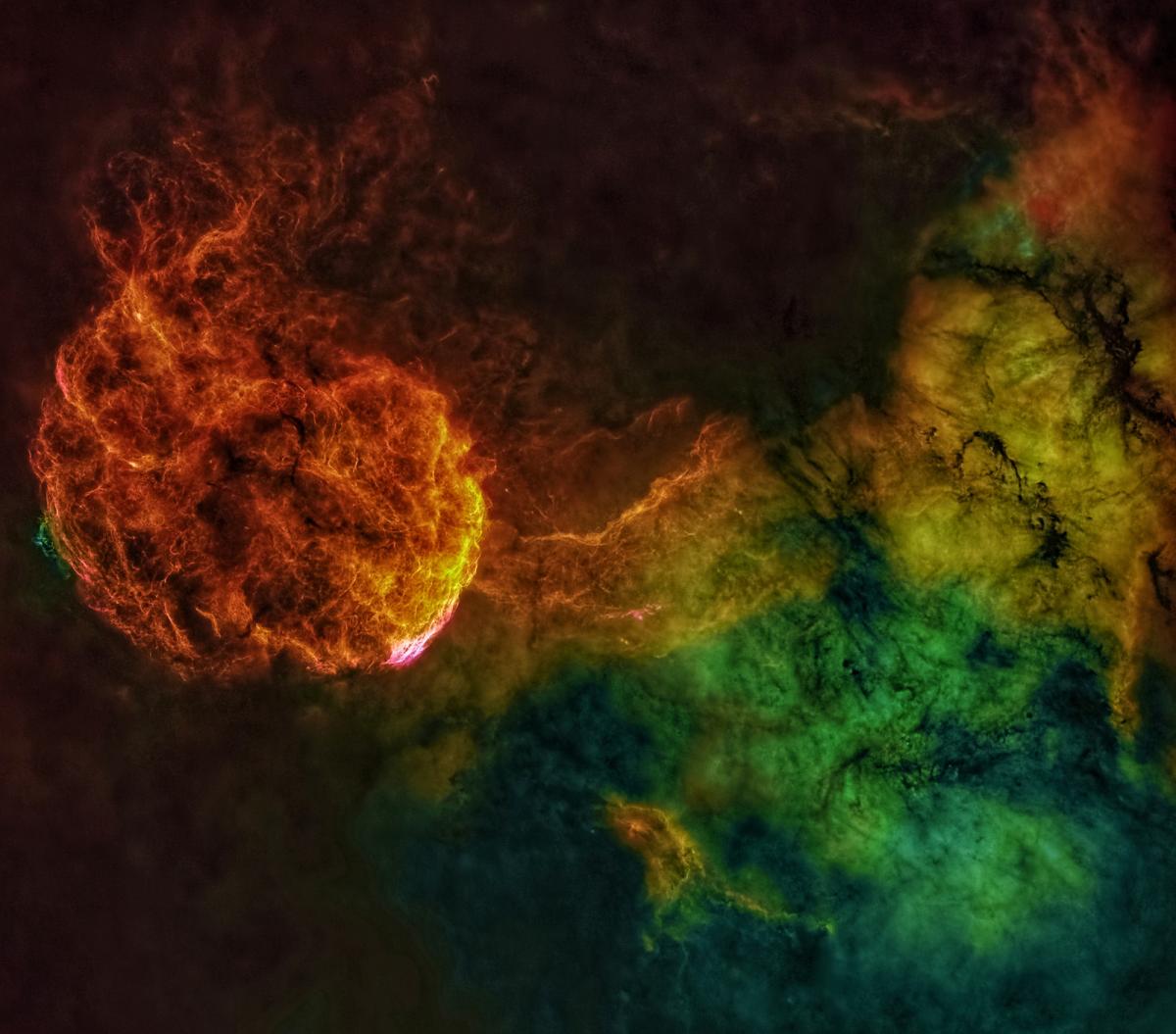 Image of a nebula which resembles a jellyfish on the left in oranges and yellows, with a green cloud on the right hand side