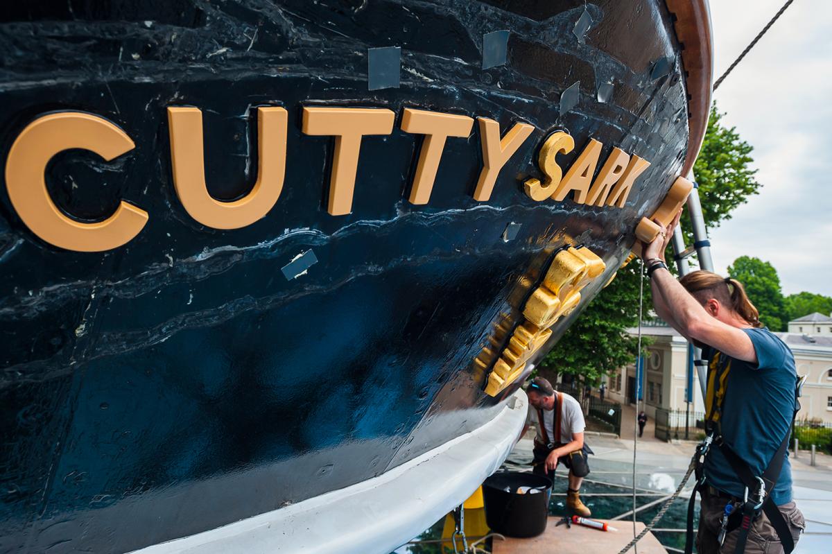 A man in a harness works on historic ship Cutty Sark, with the name of the ship picked out in gold lettering