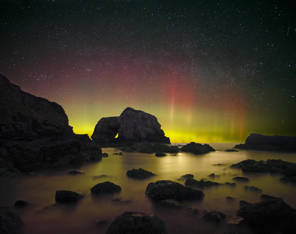 Image showing a big rocky sea arch with a thick fog lying over the sea in the foreground. In the background is a very starry black sky, with an almost neon yellowy-green aurora close to the horizon