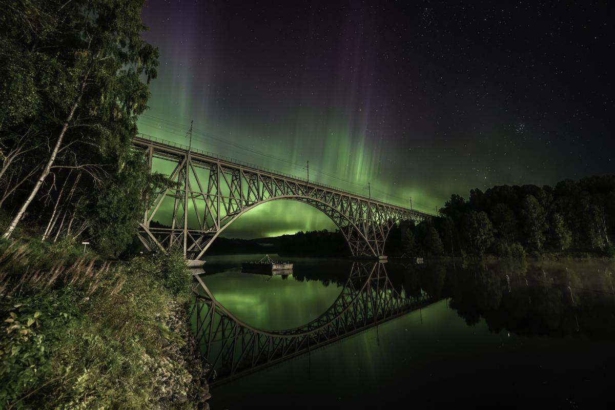 Image of a suspension-type bridge over a still, dark body of water with a landscape covered by trees in the distance. In the sky is a bright and vivid green aurora which almost forms spikes at the top. The aurora is reflected in the water, making it glow green