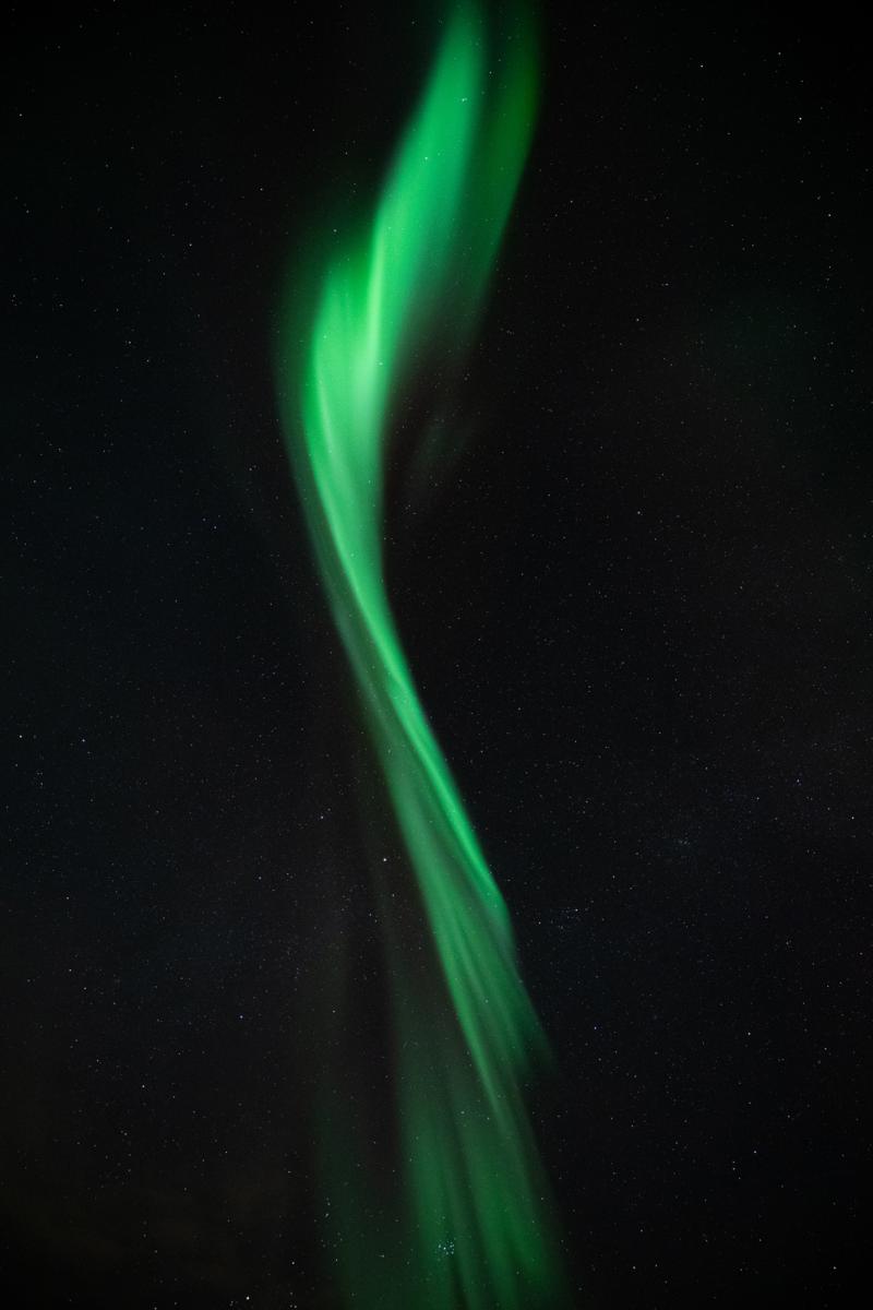 Image of the night sky in portrait form, with aurorae forming what looks like a green brush stroke right down the centre of the image, curving and twisting as it moves downwards