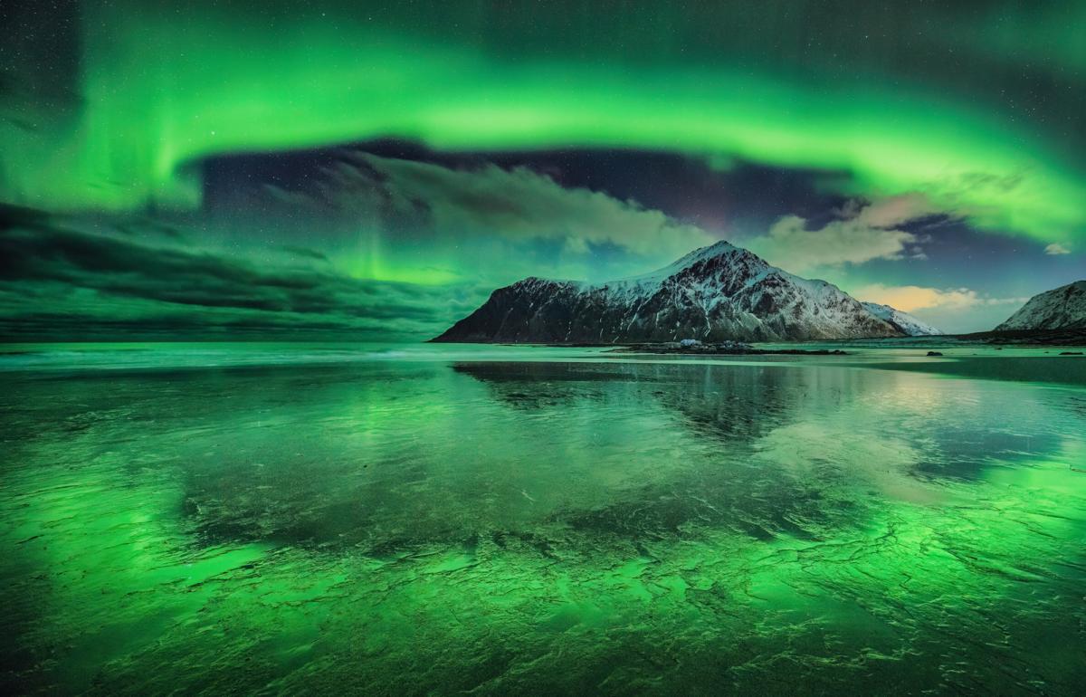 Image of a snow-capped mountain next to a wide expanse of frozen water, with a vivid aurora making almost an arc in the sky from left to right, which is reflected in the ice below