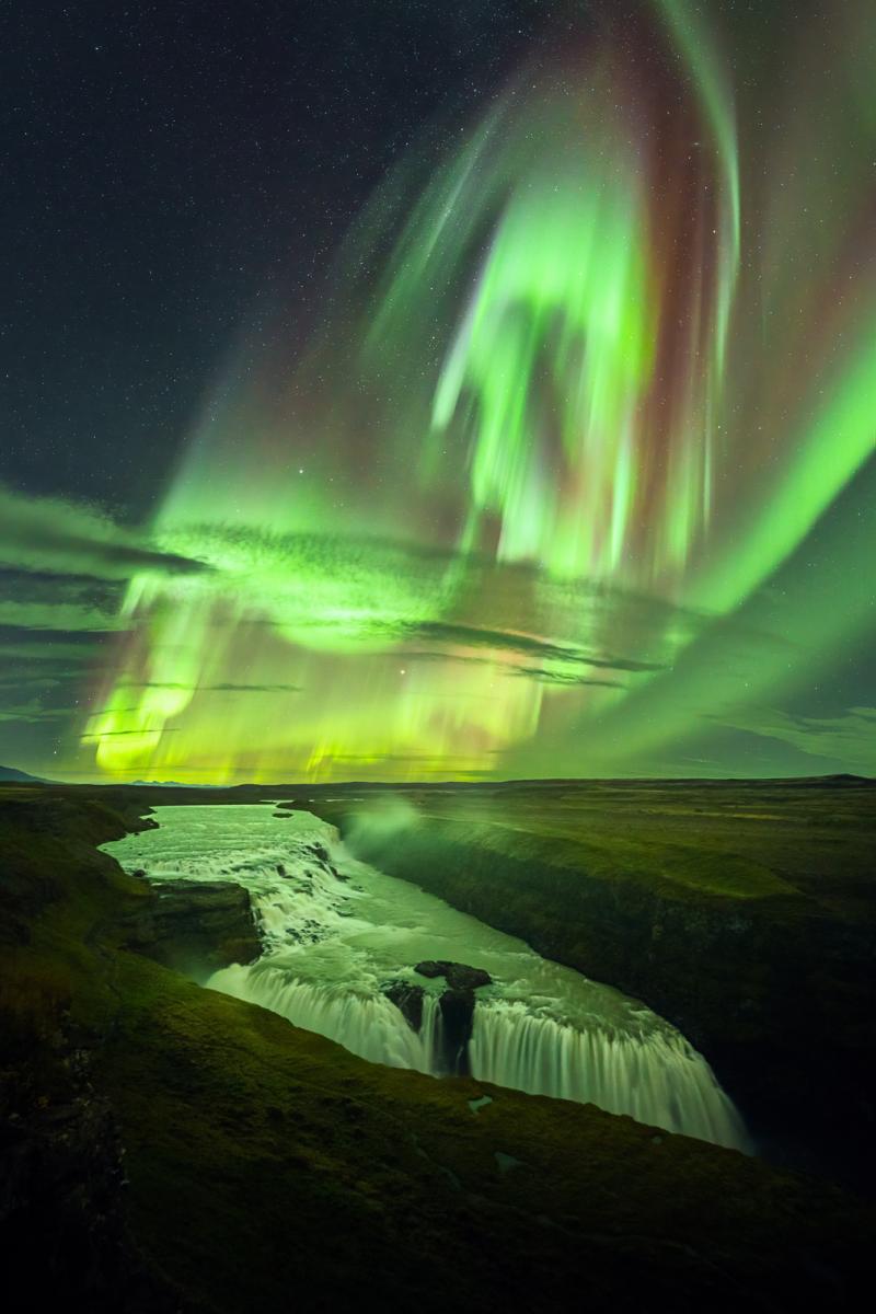 Image of a waterfall dropping into a rock formation, with the vivid aurorae in the sky behind, in many different shades of greens and different shapes which reflect onto the waterfall below