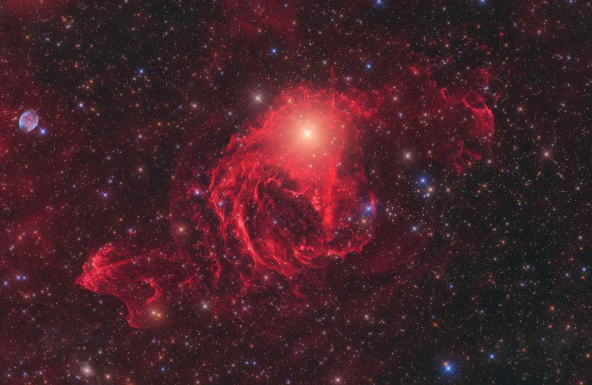 Image showing vivid red nebula in rippling waves, with a bright yellow star at the centre of the image, against a starry sky filled with blue, orange and white stars