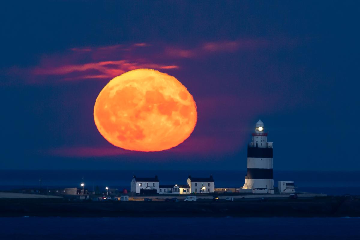 Large orange full moon in an indigo sky stretches over a black and white striped lighthouse