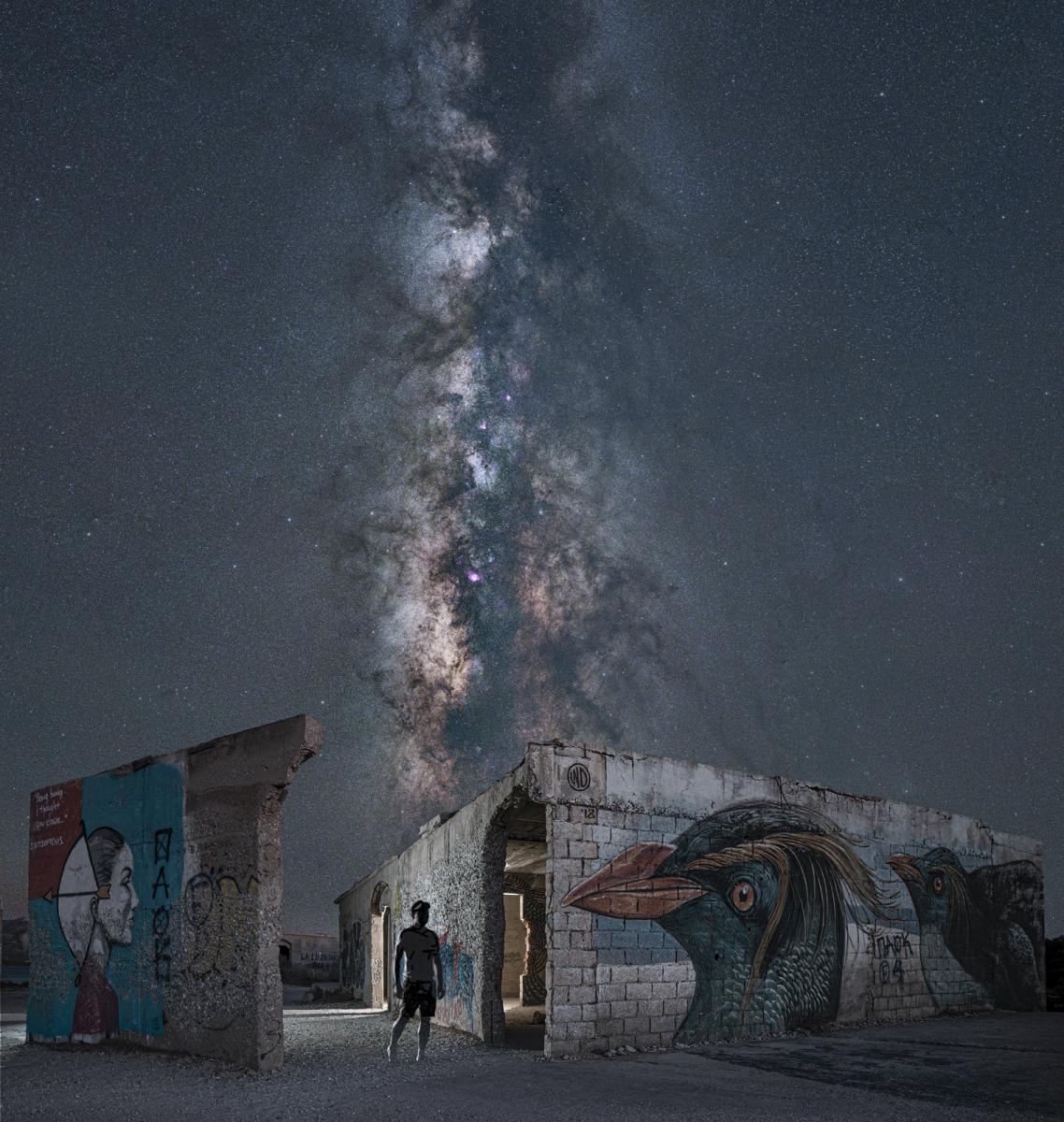 A night sky photograph of the Milky Way taken amongst ruined buildings. The eerie scene features graffiti on crumbling walls, with the figure of a man in shadow standing by a painting of a bird. A stream of stars bisects the sky above them