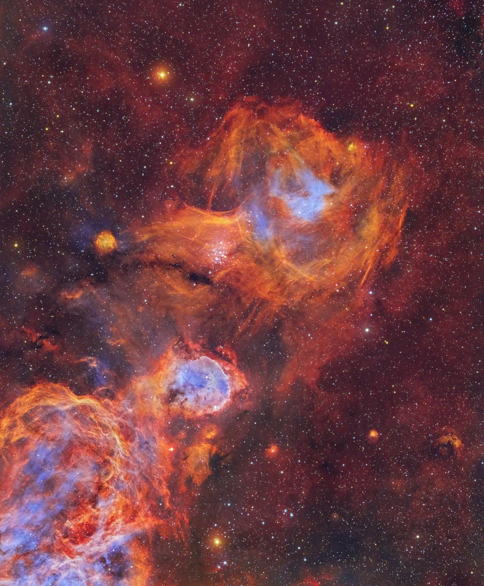 Image showing vivid nebulae in hot oranges and reds and cool blues, resembling a flame. The background is a starry sky coloured in dark reds with hundreds of stars