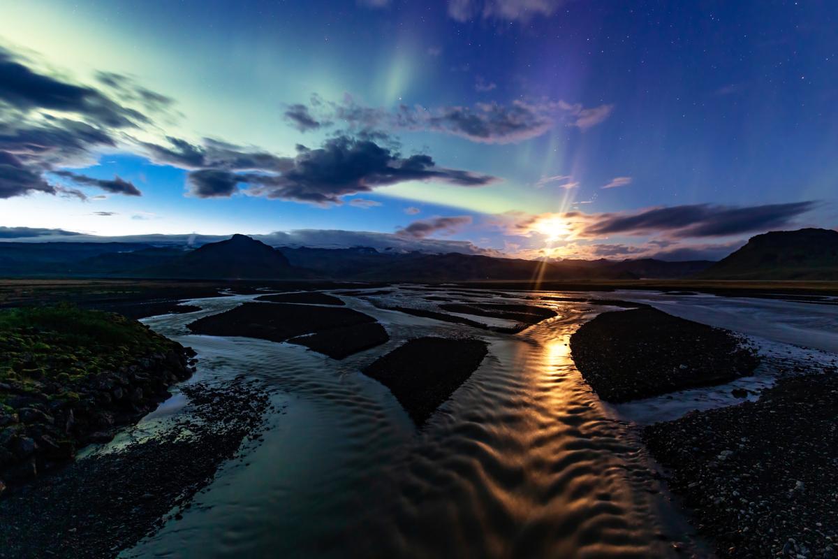 Image of an almost-frozen stream in a cold country with the dusk sky above featuring some clouds and a long trail of light green aurorae. The setting Sun is just about visible behind a cloud, lightening up the sky