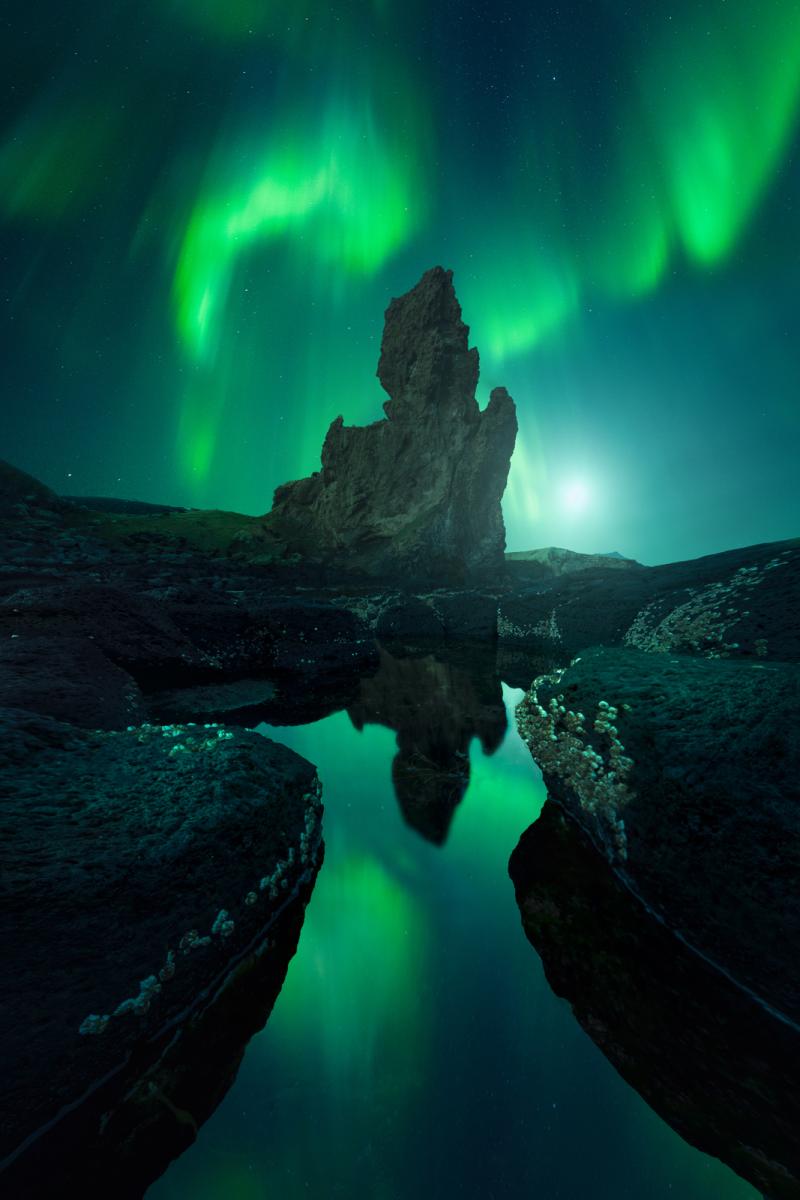 Astrophotograph of a unique rock formation within a pool of water, with the aurorae in the night sky, in bright shades of green which are reflected in the water