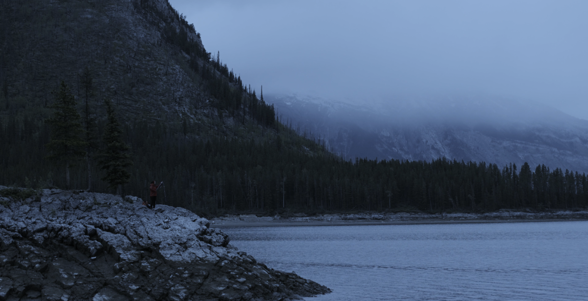 Image of cold landscape with a lake and mountains in the background. A person wearing a red coat stands next to a tripod with a camera on top of it