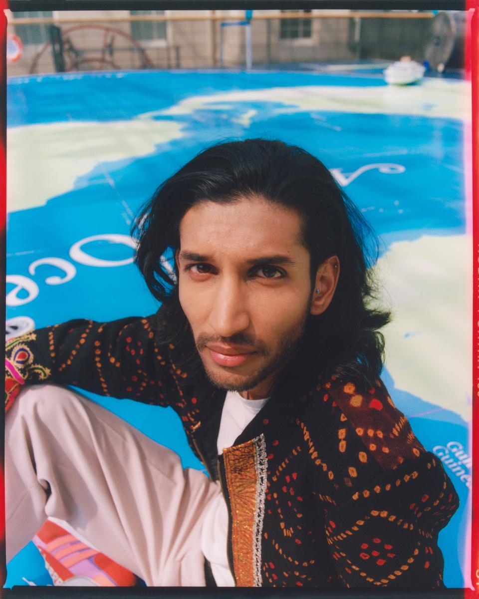 Portrait photograph of a young man with brown skin, a closely trimmed black beard and long black hair. He is sitting on the ground, his face turned upwards towards the camera