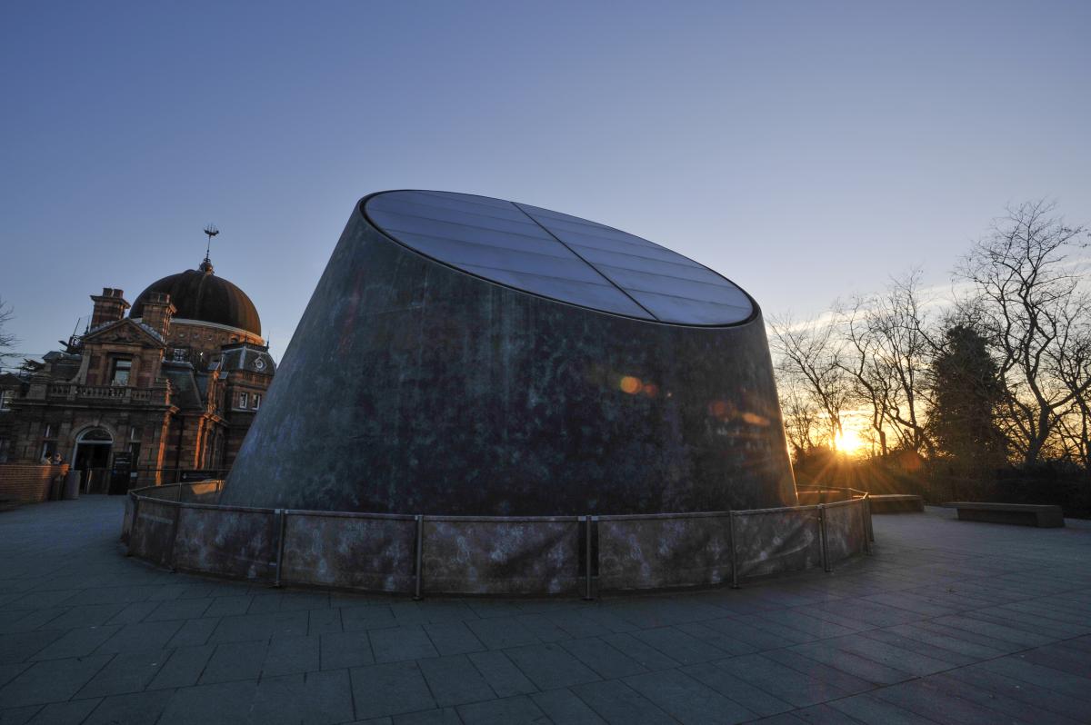 The grey metallic dome of the Peter Harrison Planetarium, with the historic buildings of the Royal Observatory Greenwich behind. A low sun is seen just peeping through the trees in the background