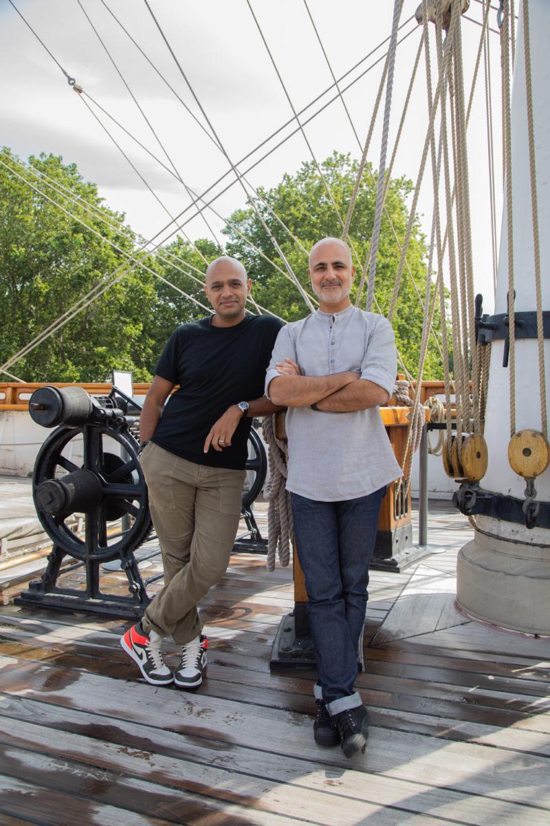 Two men stand relaxed on the main deck of an historic ship. Riggin and a mast are in the background, and the pair are smiling and looking directly at the camera