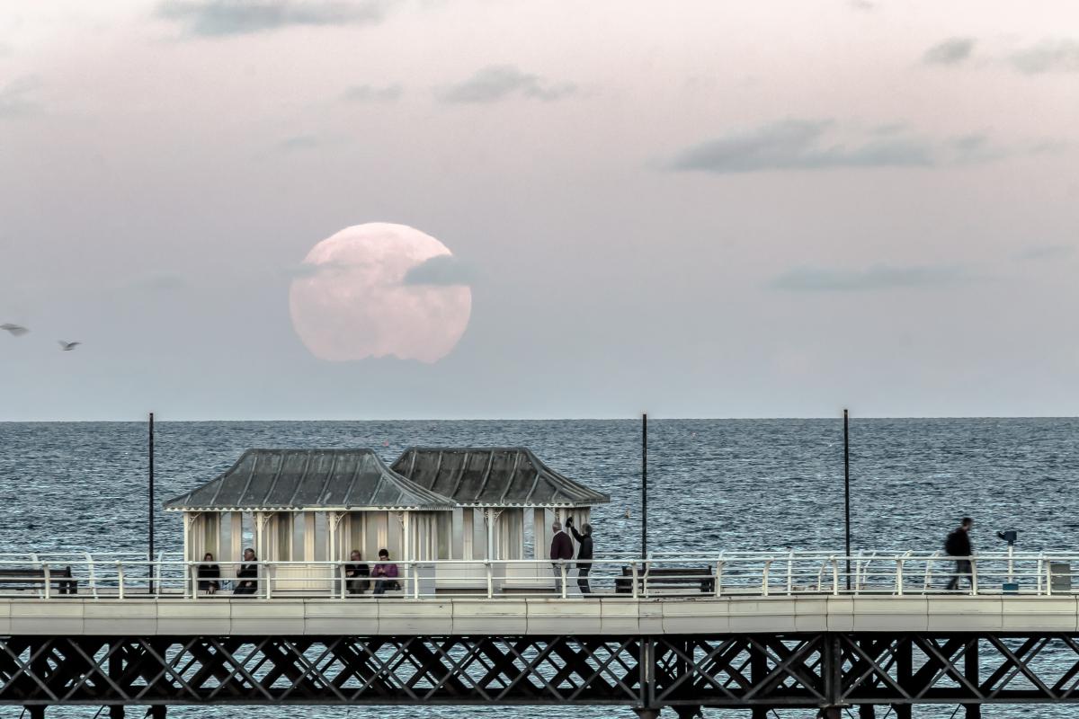 Image of the end of a pier with the ocean behind, with a large supermoon rising above the horizon in the background