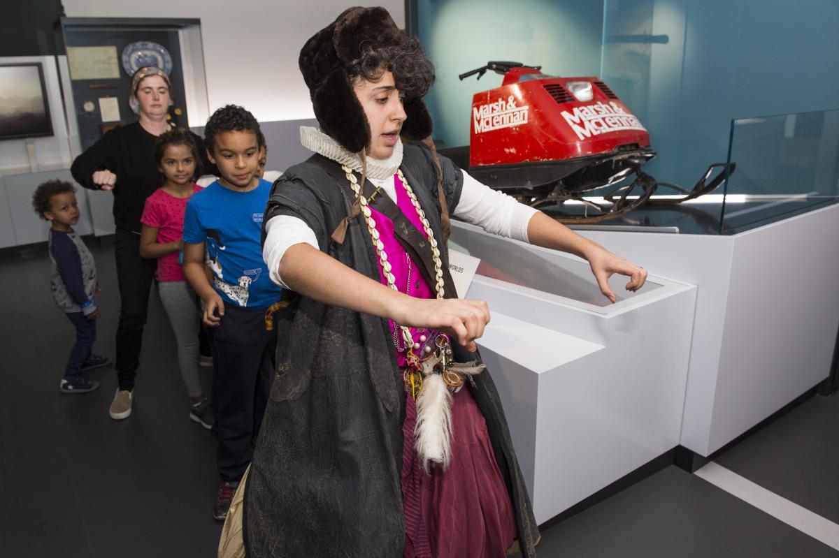 A character actor encourages a group of visitors to mimic riding a skidoo.