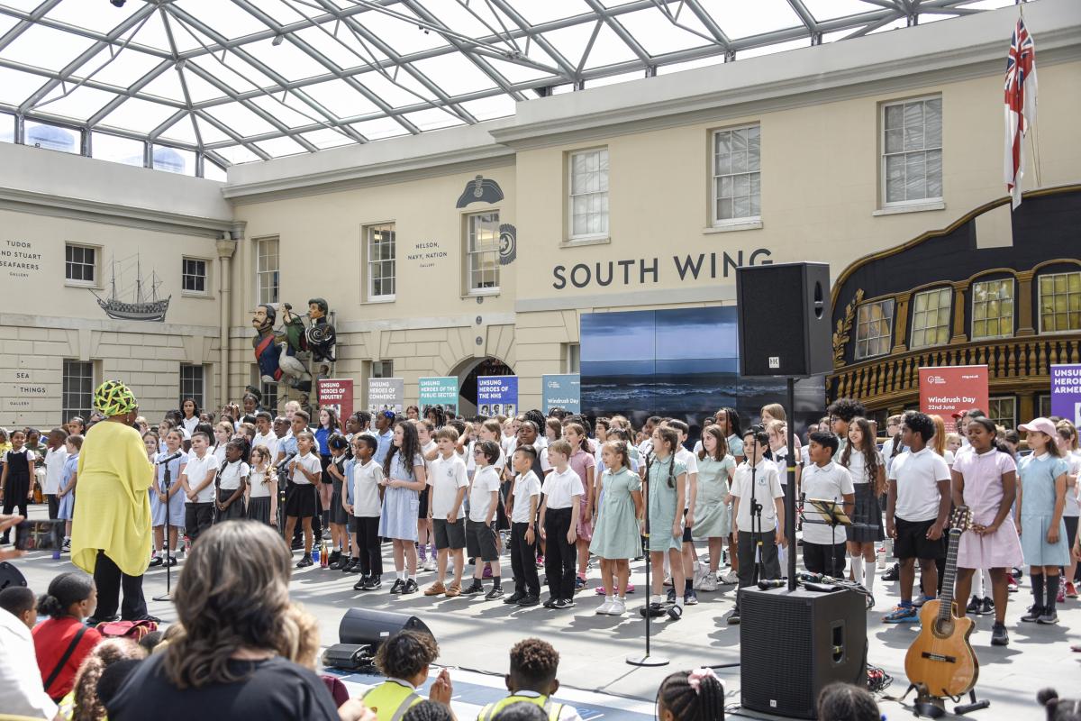 Image of choir of children standing inside a building with a big glass roof being led by a woman wearing yellow