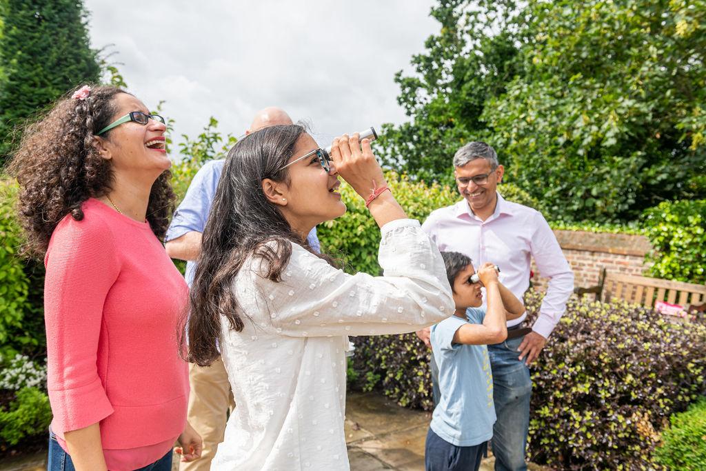 Image of a family standing in an area surrounded by greenery, with two children holding small handheld cylinders up to their faces