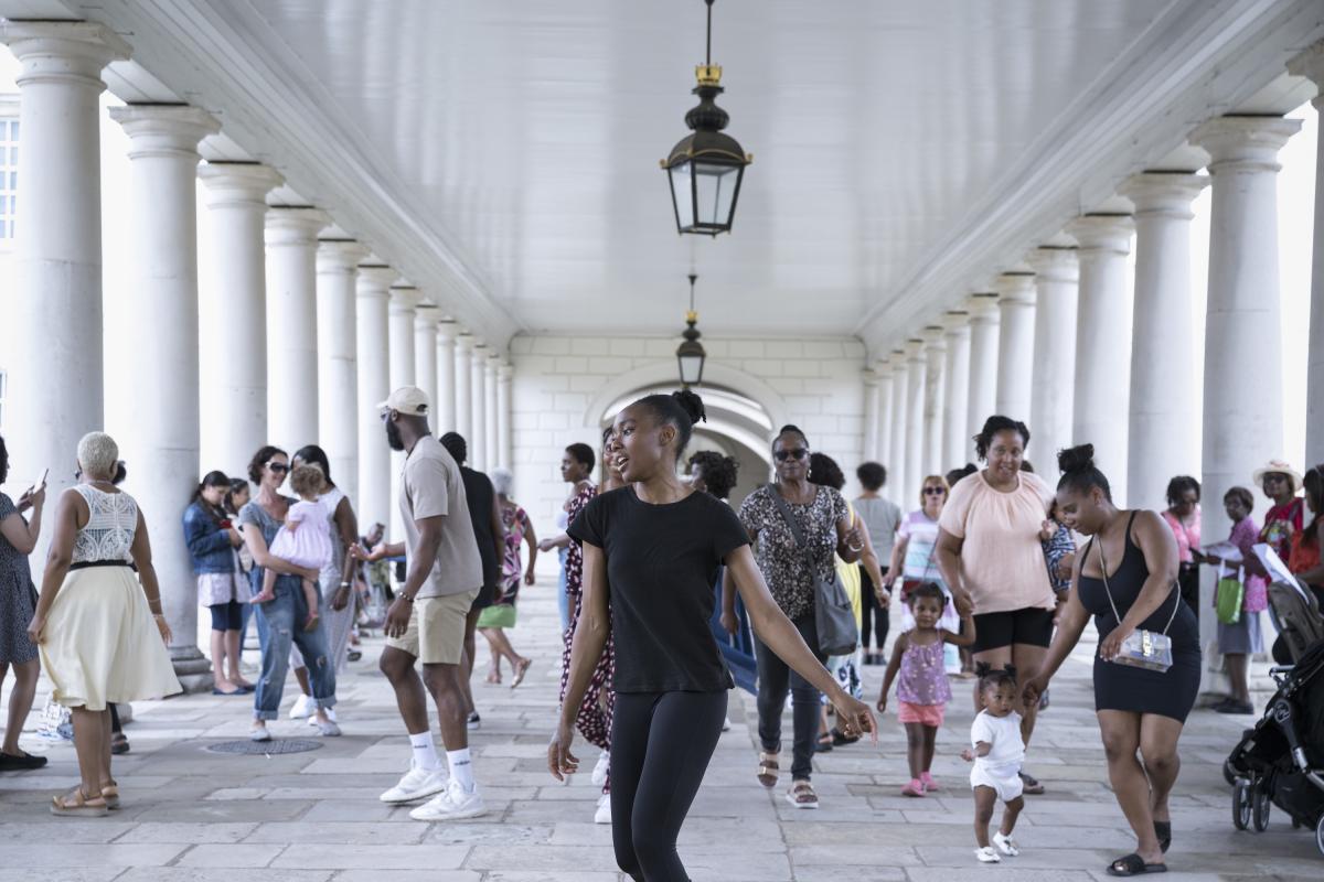 A woman dressed in black stands at the front of a group of people, framed by evenly spaced white columns and paved stone floor