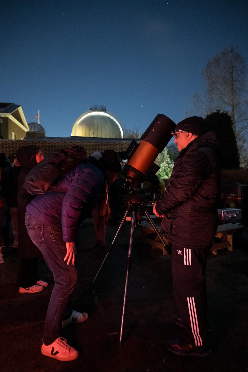 A man crouches to look through a telescope outdoors at night, with another man looking on