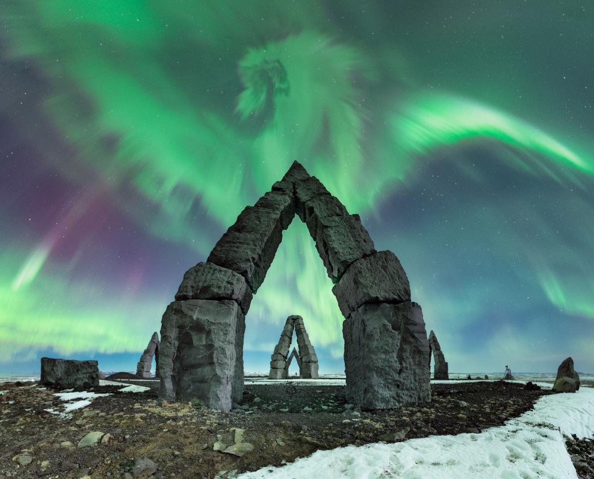 Image of multiple stone formations in arch shapes. Behind them in the sky is the aurorae in aqua greens, purples and blues. The aurora in the middle of the image resembles a dragon