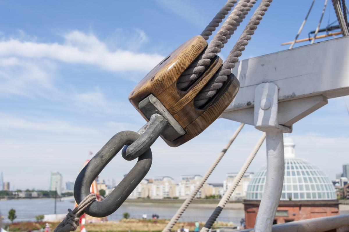 Image of a 'block' a piece of wood where pieces of rope are fed through in a ship's rigging. It's attached to a chain link and ropes are coming out of the other end. In the background is a river and a blue sky