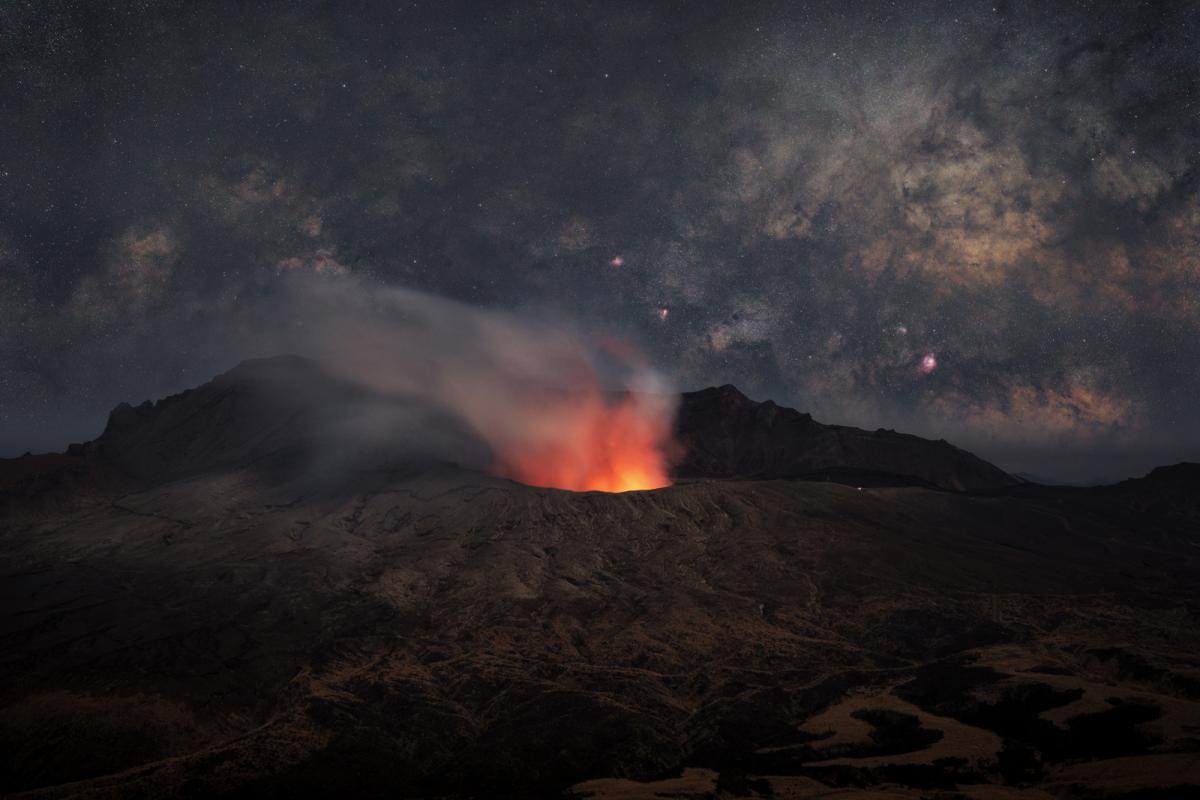 The bottom half of the image is of a volcano which is erupting with lava, around the top of the volcano are streams of dried lava and ash. In the sky behind the Milky Way is almost horizontal with patches of dark and light