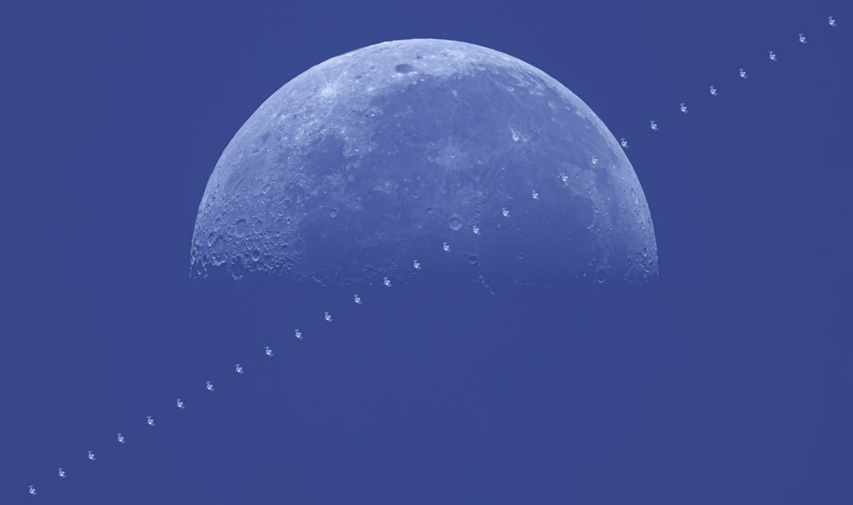 Image of half of a full Moon with a blue sky behind it. Across it diagonally is a series of dark 'H' shapes which shows the passing of the International Space Station. 