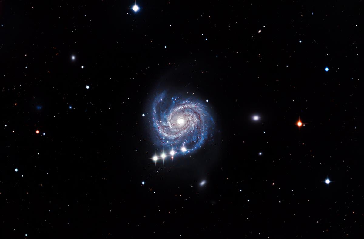 Image of a small spiral galaxy in whites, blues and purples against a black starry sky. There are four white dots crossing over at the very bottom of the galaxy, which are the dwarf planet Ceres