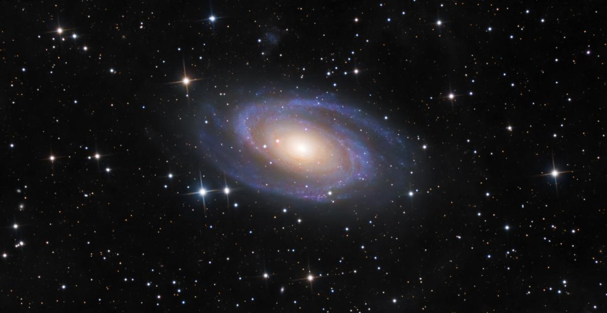 Image of a spiral galaxy in blues, whites and purples. The galaxy is at a tiled angle to us. Behind it is a black sky with stars dotted around the image.