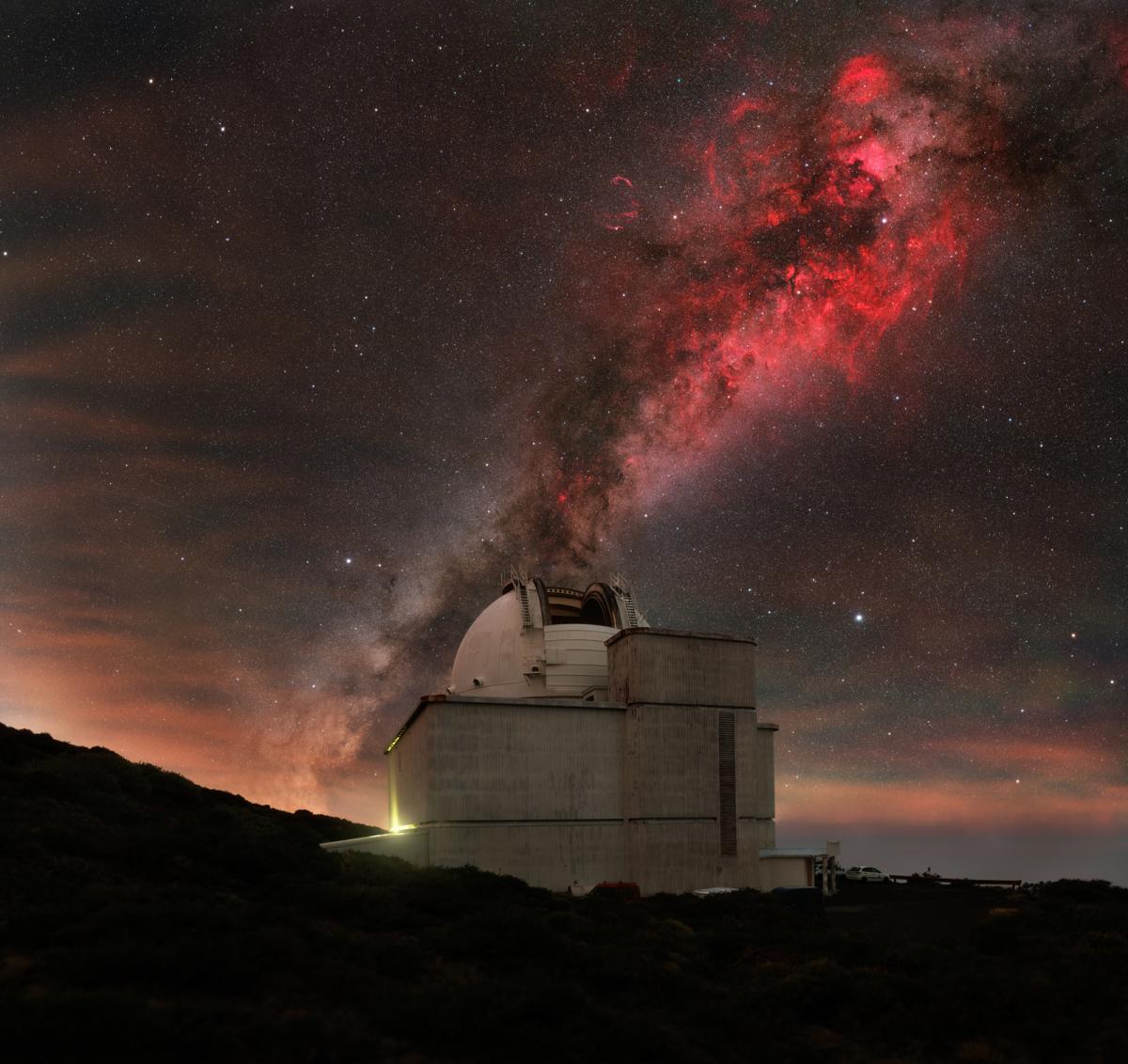 Image of an observatory with a big domed roof and behind it diagonally is a red nebula against a grey sky with hints of orange at the bottom.