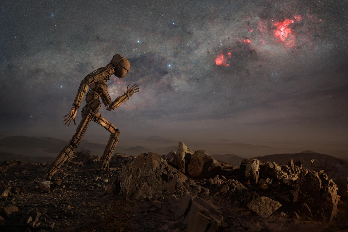 Image of a small figure made of wire and stones facing to the right in a running motion, with a dark starry sky and the Carina Nebula, bright vivid clouds of red, standing out in the sky